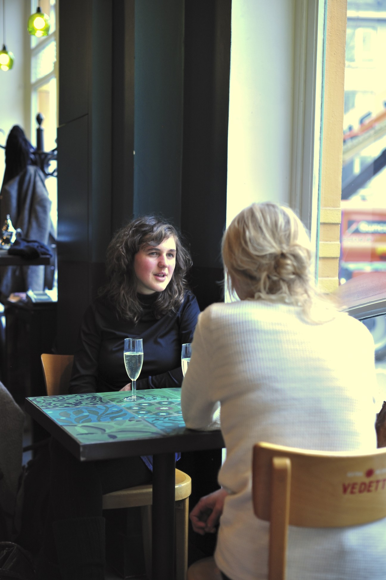 Two women conversing at a table with drinks in a modern café interior with large windows.