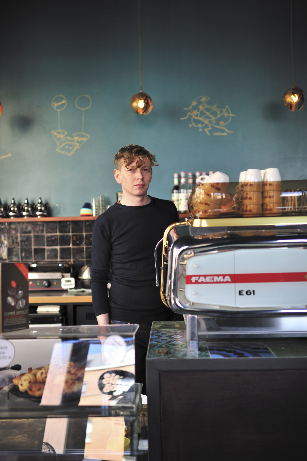 Young man in black sweater standing behind espresso machine in modern café with teal wall and hanging pendant lights.