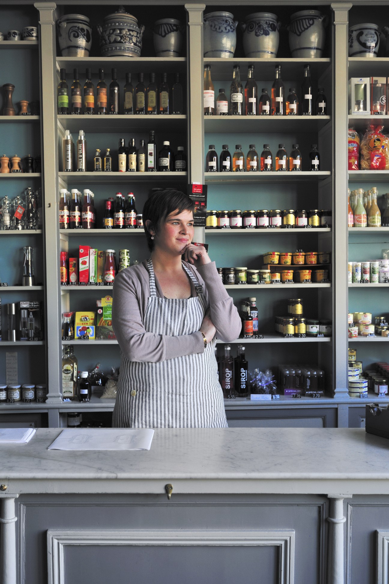 Evelyne Huughe stands behind a counter at Tierenteyn-Verlent mustard shop, with shelves of jars and bottles displayed behind her.