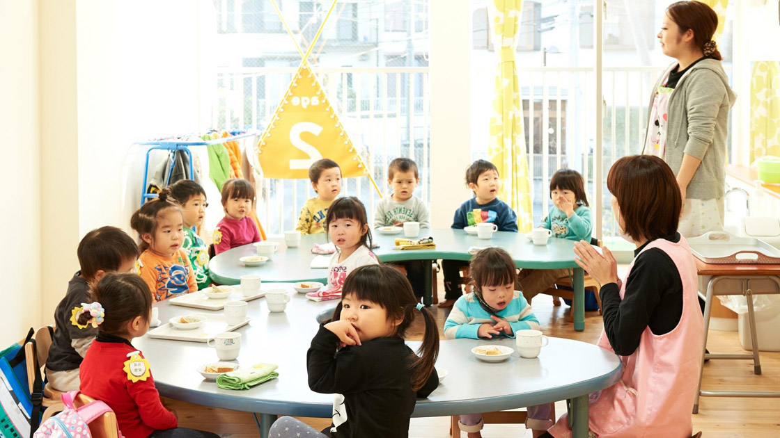 Young children seated at tables during snack time at Byobugaura Harukaze Nursery with caregiver supervising.