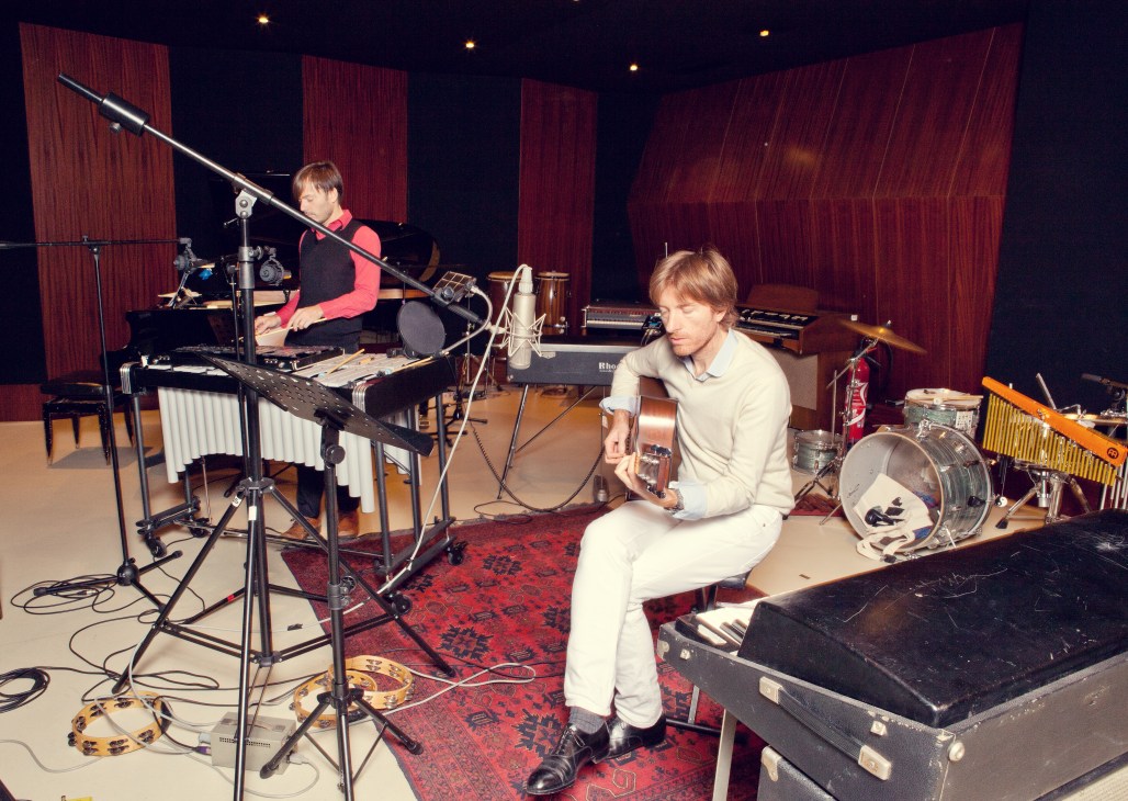 Two musicians in a recording studio surrounded by acoustic and electronic instruments, with red wooden acoustic panels.