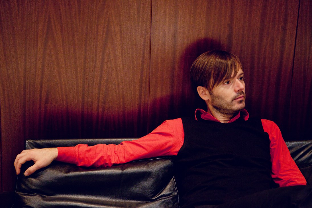 Jean-Benoît Dunckel from Air relaxes on a black leather couch against wooden walls wearing a red jacket.
