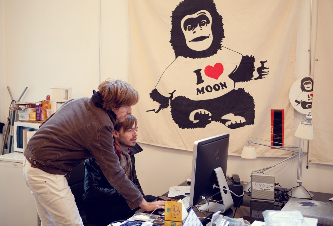 Two men work at a desk with a computer while a gorilla wall mural wearing an "I love moon" shirt looms behind them.