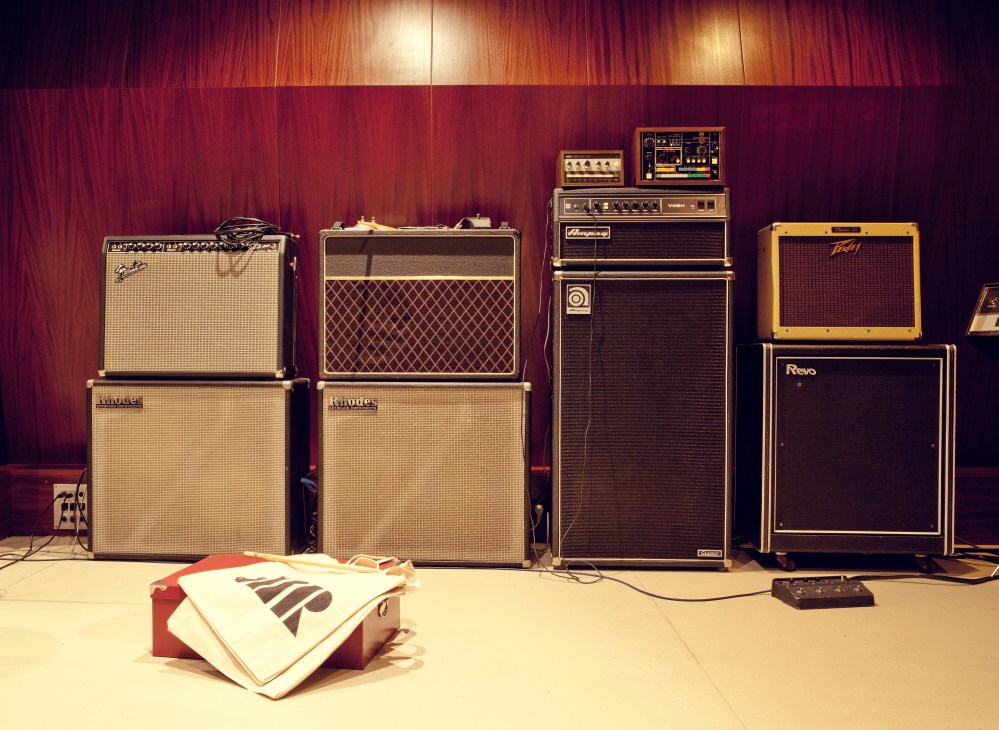 Vintage guitar amplifiers and synthesizers arranged against red wooden studio walls with warm lighting.