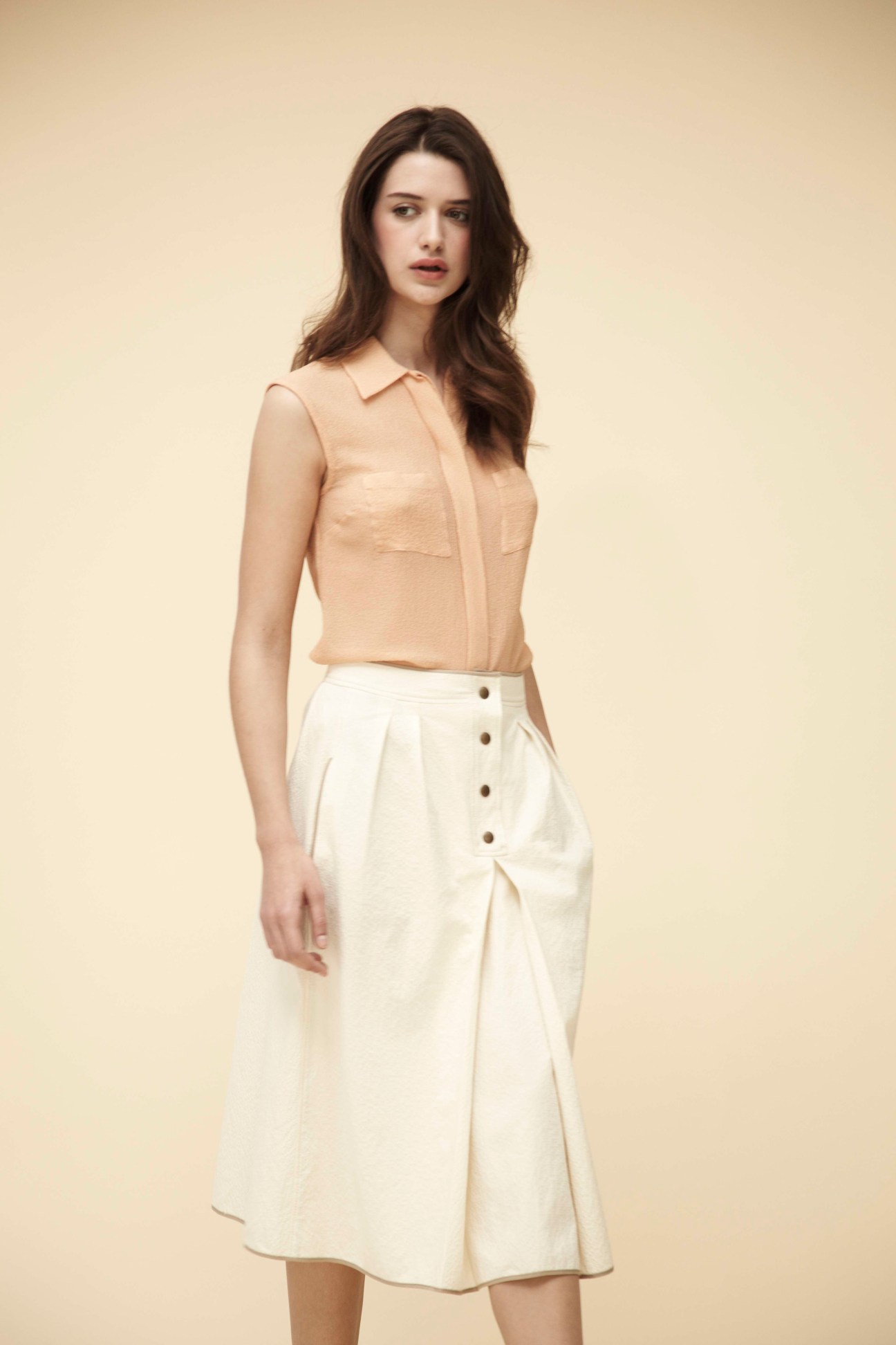 Woman wearing a sleeveless beige shirt and white button-front midi skirt against a cream background.