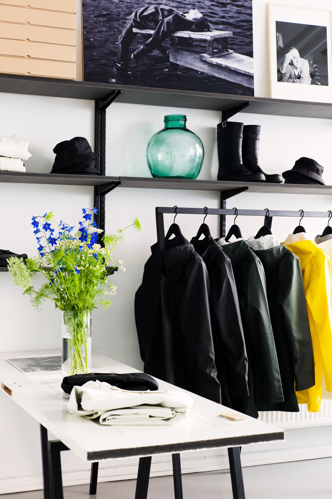 Black raincoats hanging on a rail in a retail display with shelving, flowers, and a turquoise bottle.