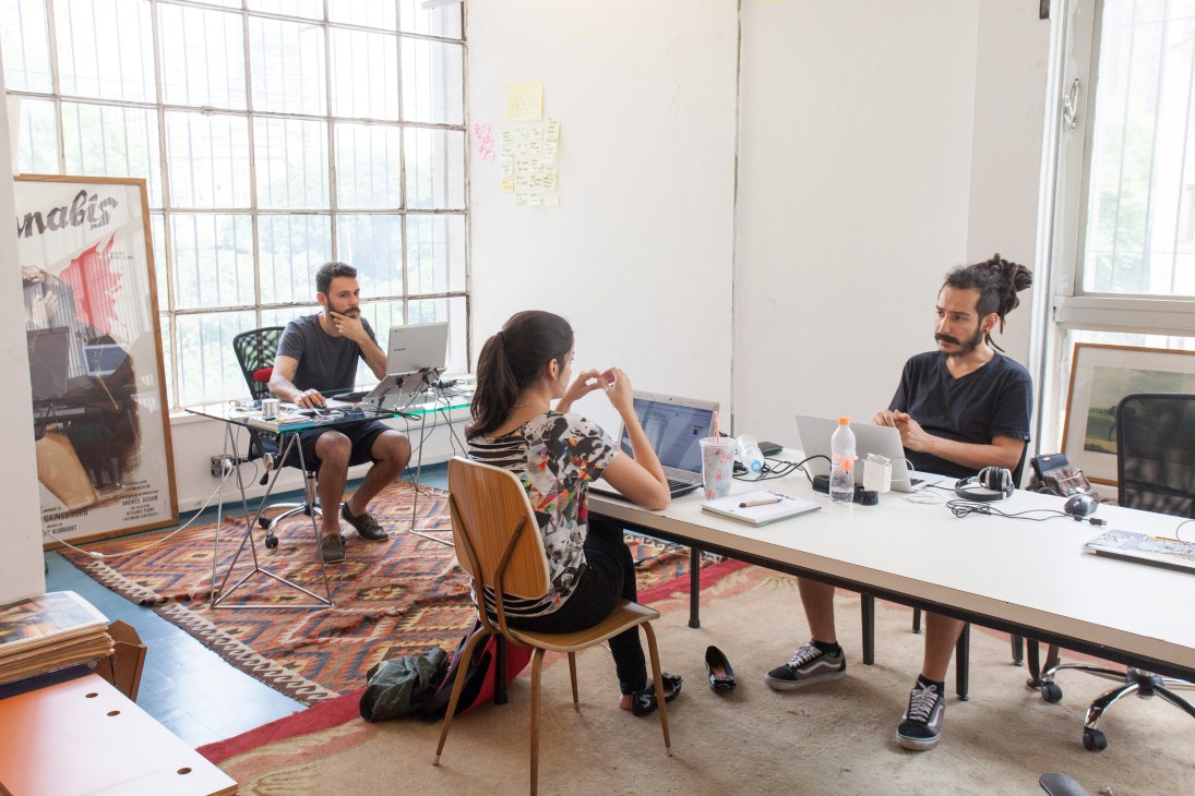 Three people work at desks in a bright newsroom with large windows and colorful rugs.