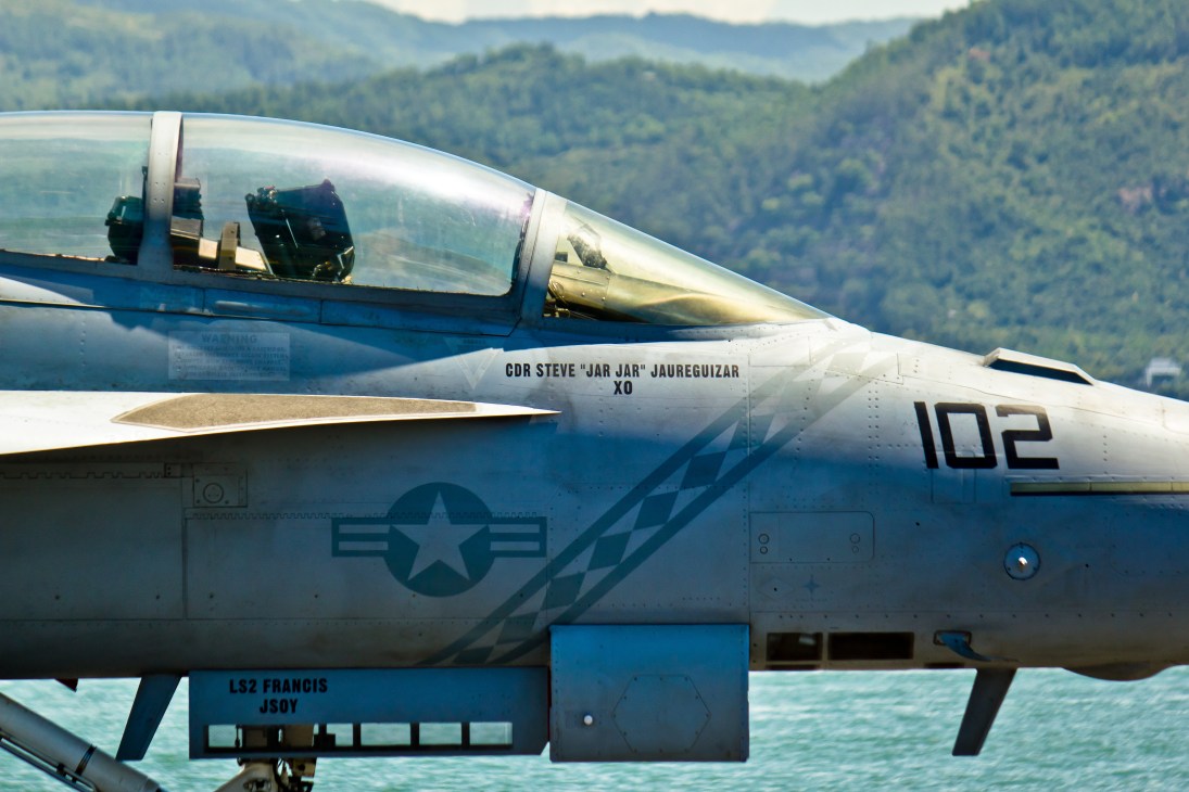 Close-up of F/A-18 Super Hornet fighter jet cockpit with pilot visible through canopy, forested mountains in background.
