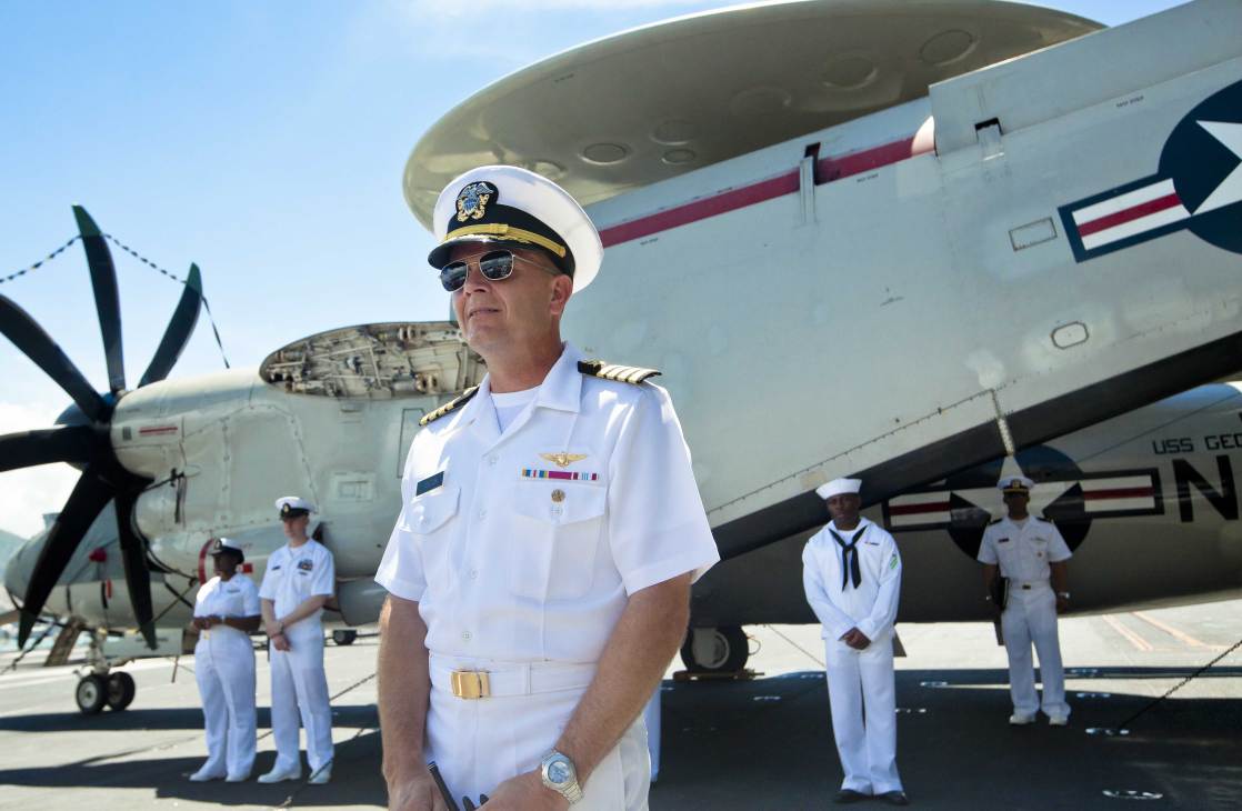 US Navy officer in white uniform stands before an E-2C Hawkeye surveillance aircraft with crew members in background.