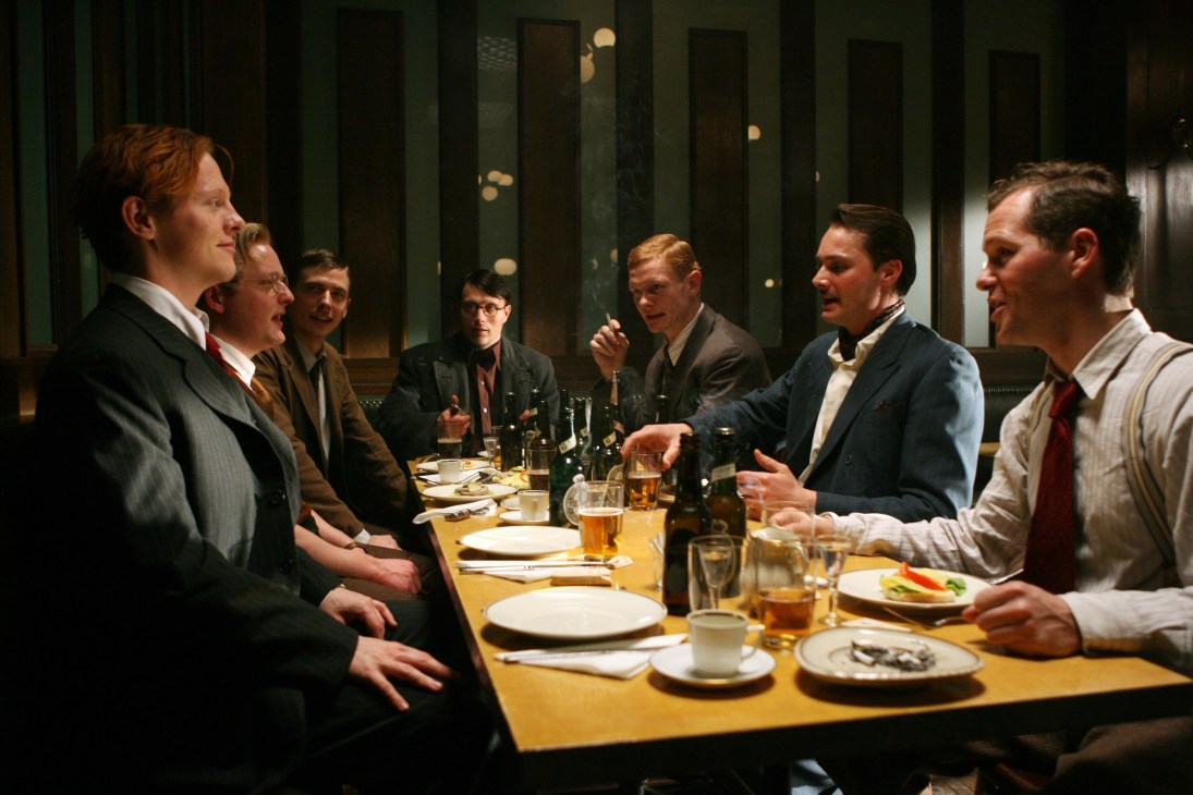 Men in suits seated at a dining table during an intense conversation in a formal interior setting.