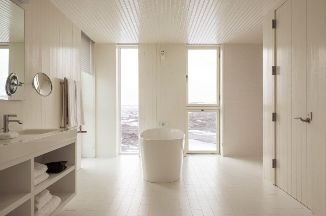 Minimalist bathroom with freestanding white bathtub, sink vanity, and ocean views through glass doors at Fogo Island Inn.