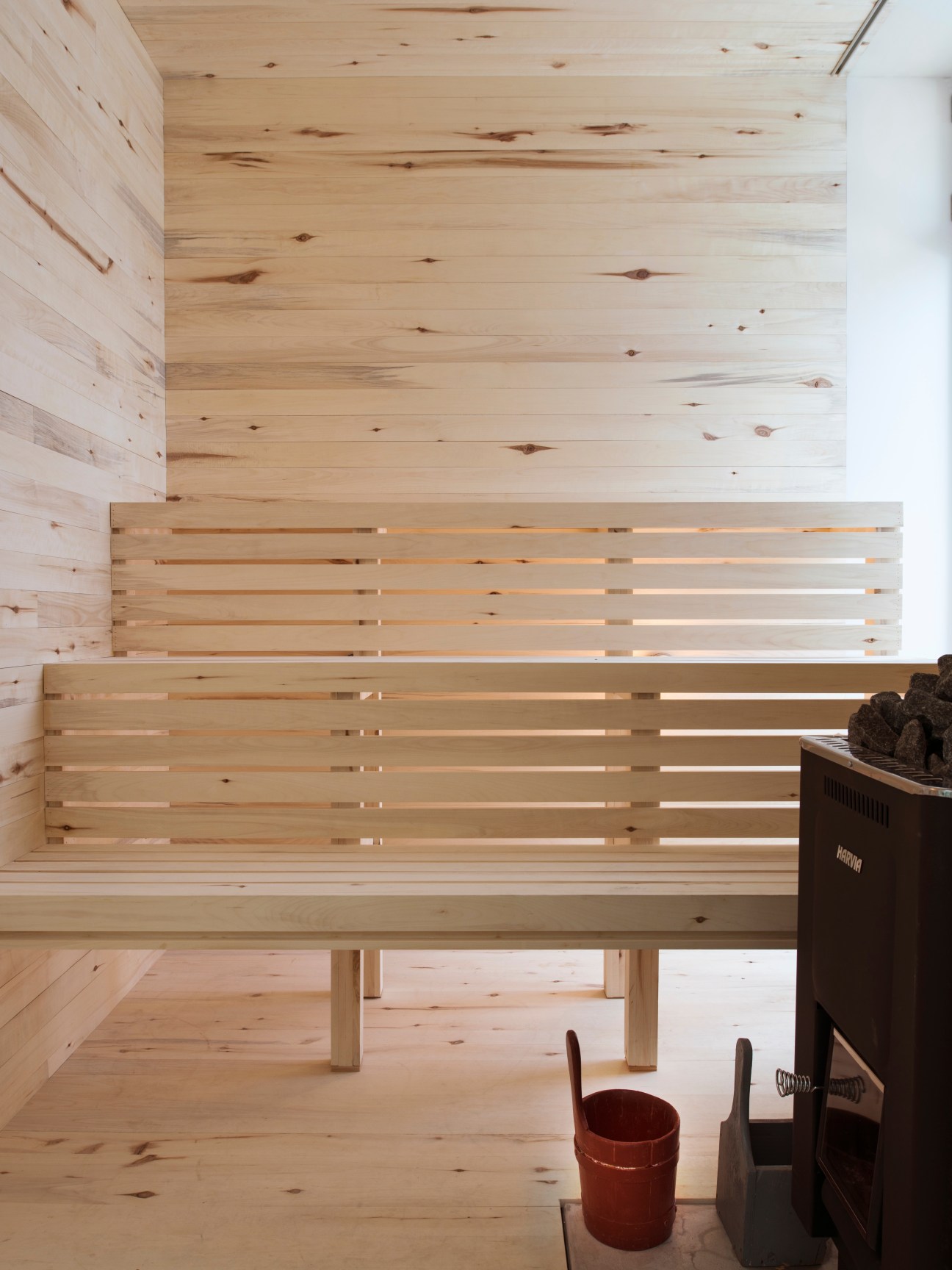Interior of a minimalist aspen wood sauna with slatted benches and ceiling, with a dark stove visible on the right.