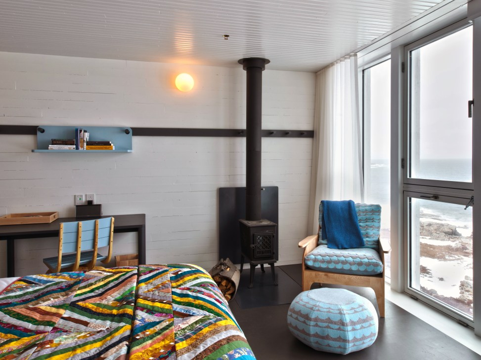 Modern bedroom with colorful quilt, wood stove, and ocean view through glass doors at Fogo Island Inn.