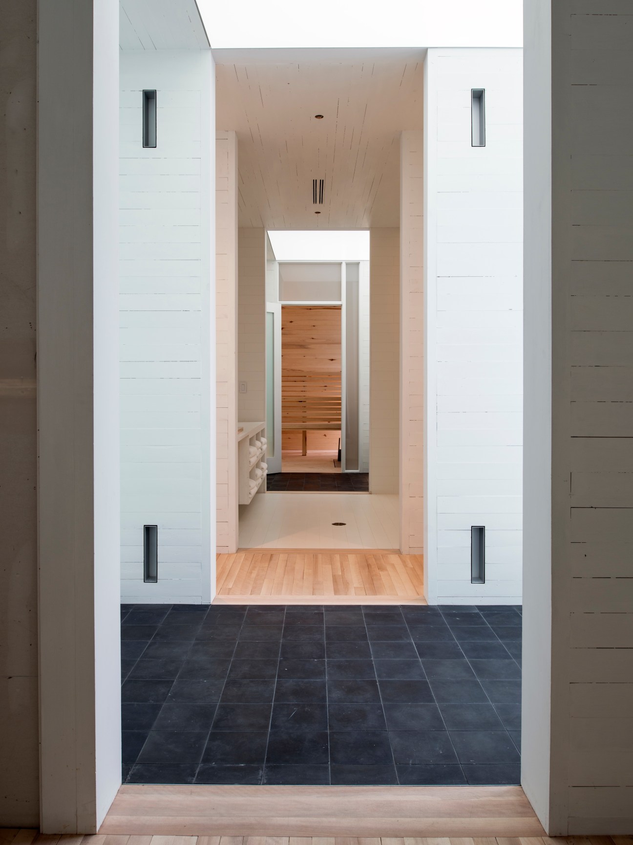 Minimalist corridor entrance to a sauna with white walls, dark tile flooring, and wooden interior visible beyond.