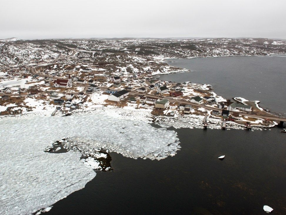 Aerial view of the town of Fogo with colorful wooden houses surrounding a harbor, surrounded by pack ice and snow-covered landscape.