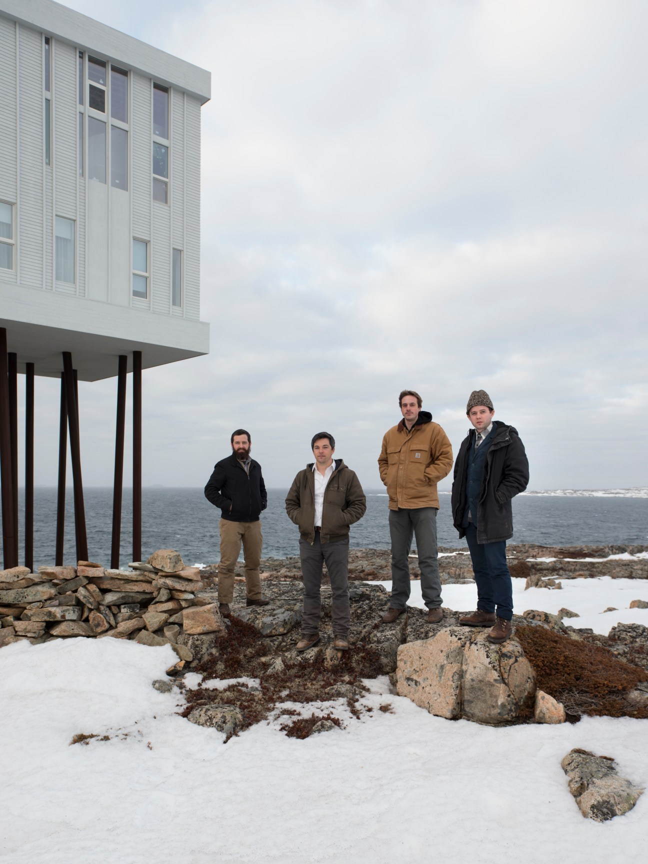 Four male architects stand on rocky snow-covered ground in front of a modern timber-clad building on stilts overlooking the ocean.