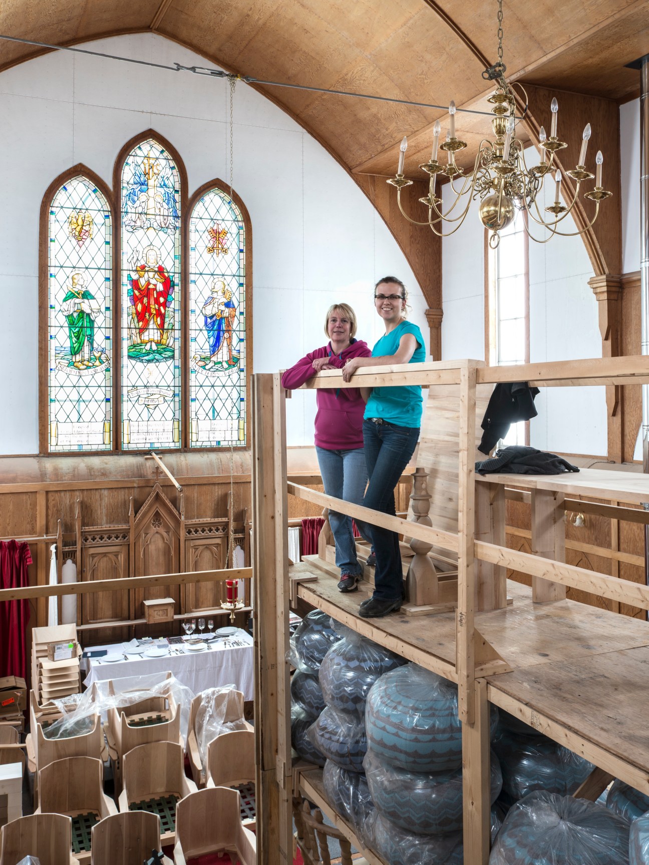 Two women standing in a deconsecrated church repurposed for storage, with stained glass windows and wooden scaffolding visible.