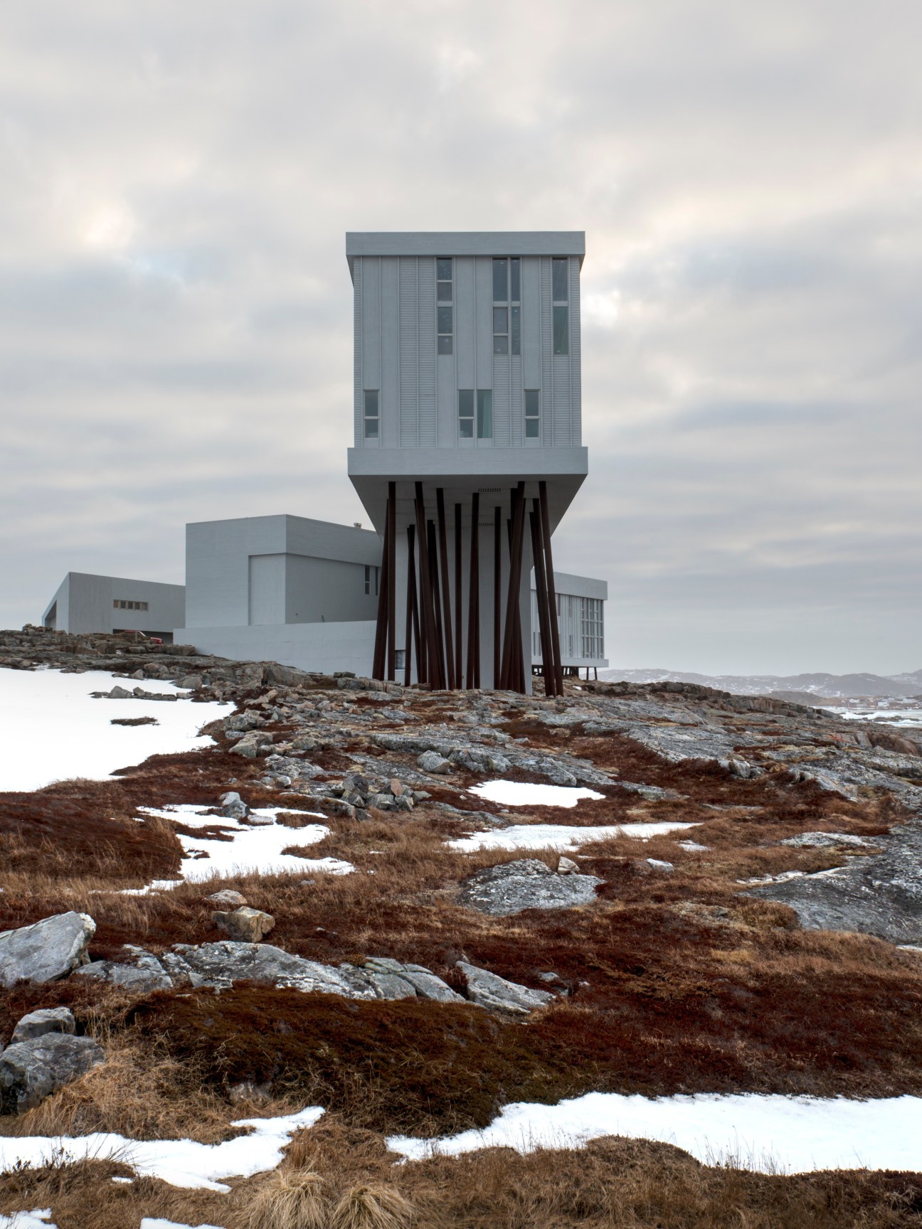 Modern timber-clad hotel building elevated on stilts overlooks rocky coastline with patches of snow.