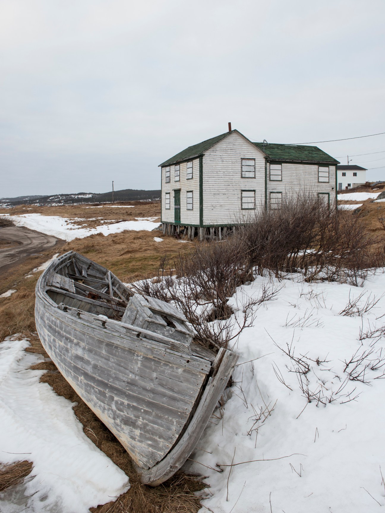 Weathered wooden boat on snowy ground with elevated wooden house and bare landscape in background.