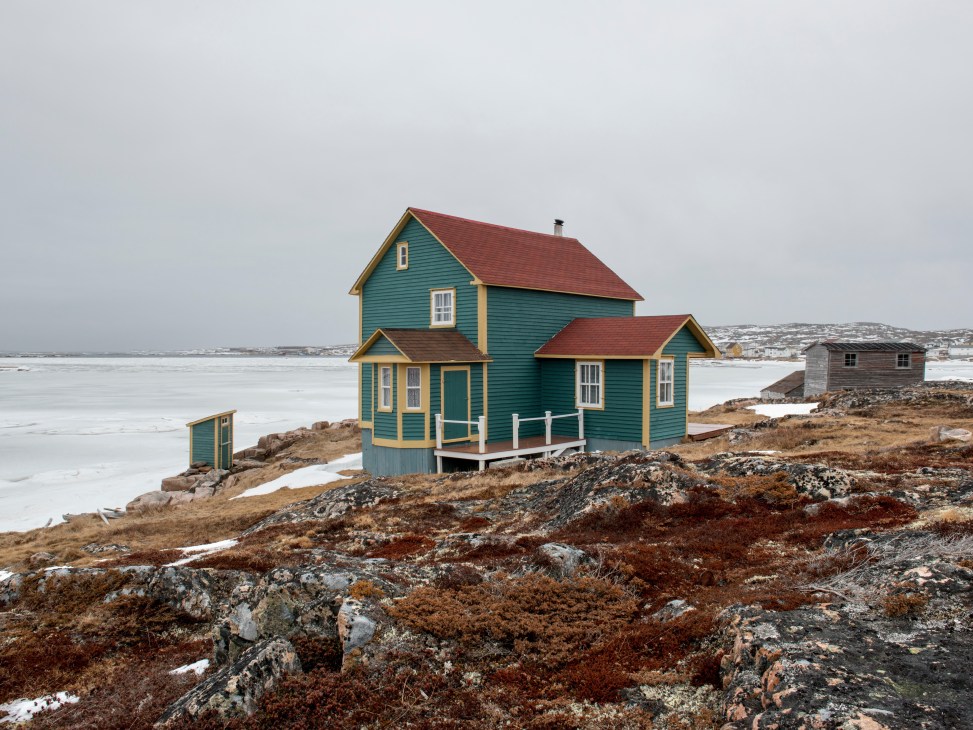 Turquoise and red wooden house with yellow trim on rocky Fogo Island coastline overlooking frozen water.