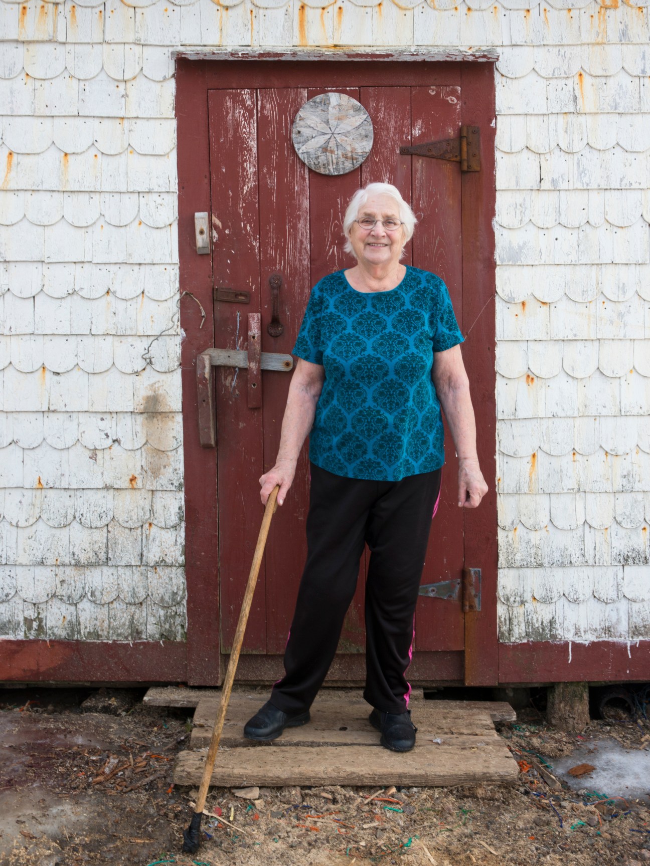 Elderly woman in blue patterned shirt standing outside white wooden shed with red door on Fogo Island.