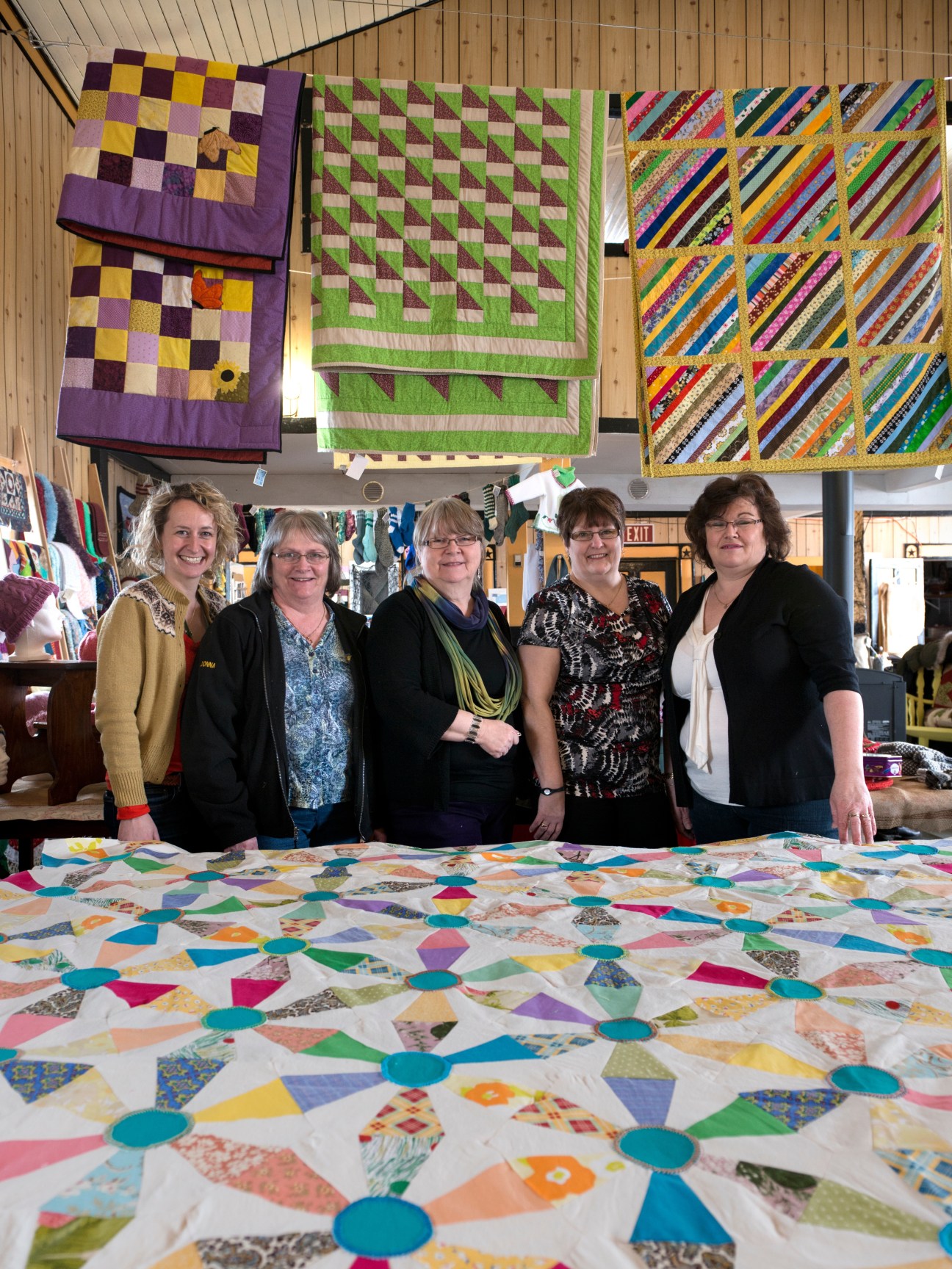 Yvonne Mullock and five women from Fogo Island's textile guild stand with their handmade quilts displayed behind them.