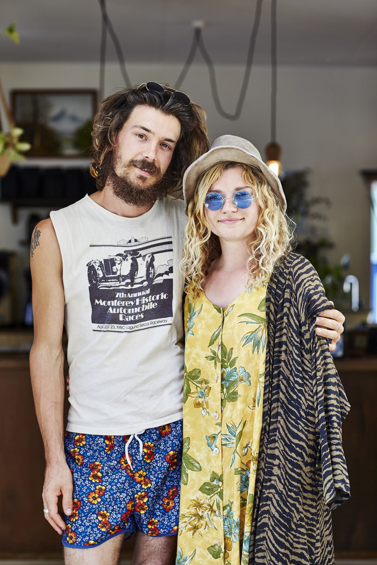 Two people posing together inside Folk Café, a man with long dark hair and beard wearing a white sleeveless shirt, and a woman with blonde wavy hair wearing blue-tinted sunglasses and a yellow floral dress.