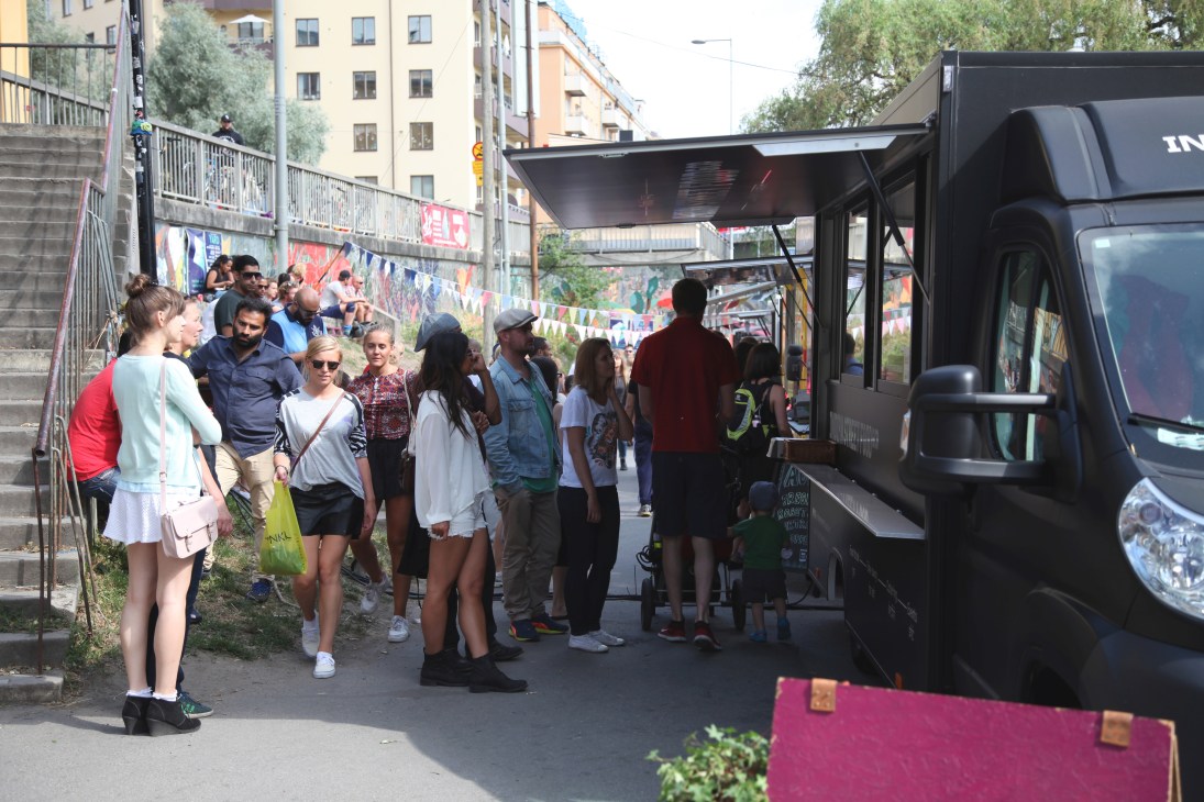 Customers queue at a black food truck in a Stockholm street with colorful murals and urban architecture.