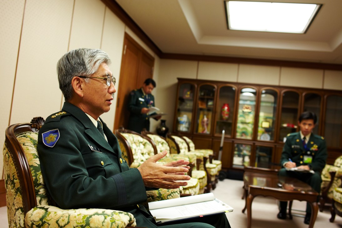 General Eiji Kimizuka in GSDF uniform gestures while speaking in a formal office meeting room.