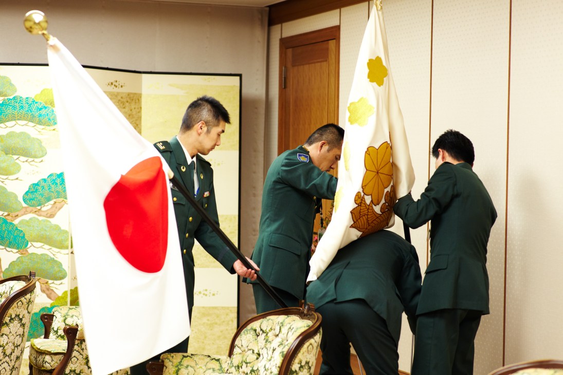 GSDF staff in military uniforms arranging the Japanese national flag and GSDF insignia flag in a meeting room.