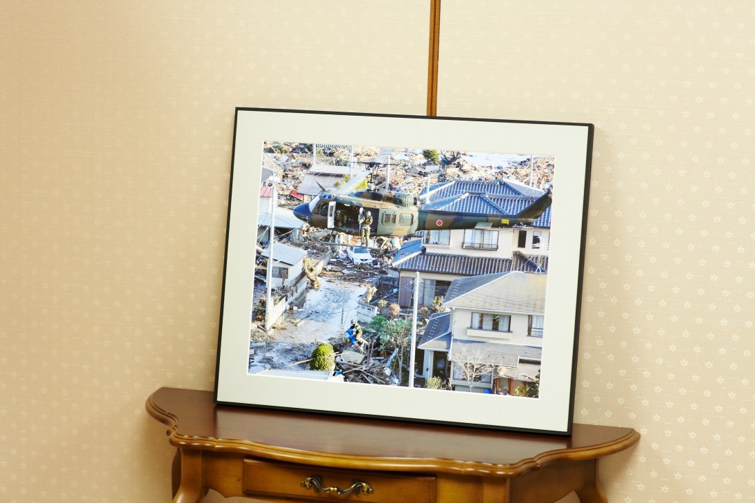 Framed photograph showing GSDF helicopter and personnel conducting tsunami rescue operations amid destroyed buildings and debris.