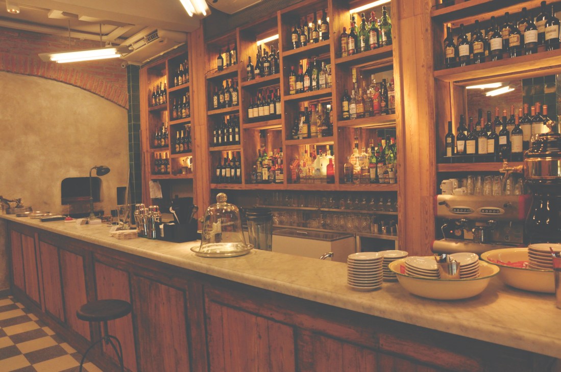 Interior of Fornería restaurant with wooden bar, shelving displaying wine bottles, and checkered floor.