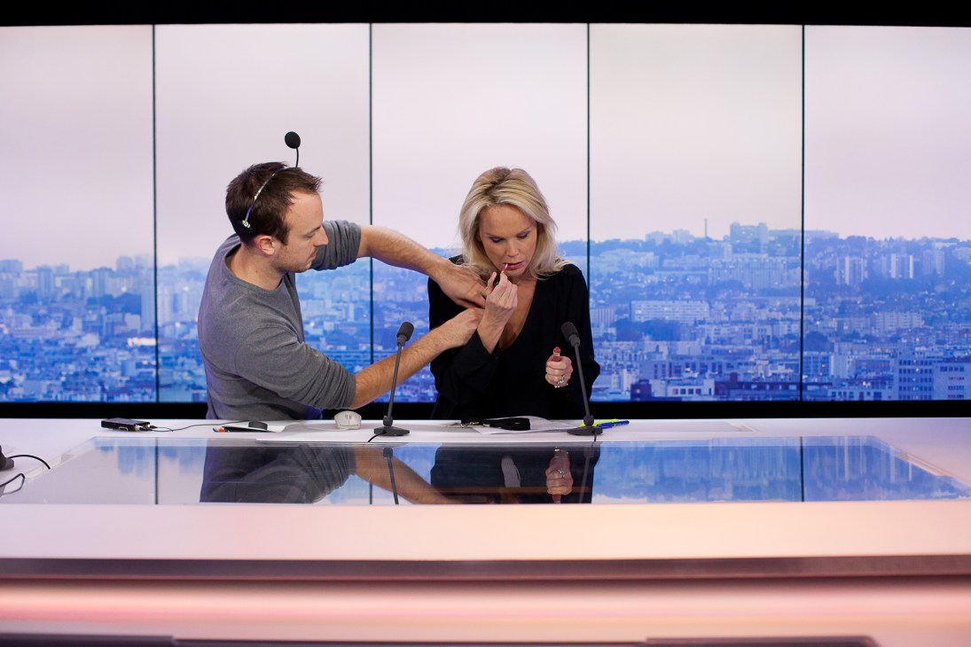 A makeup artist adjusts a female presenter's makeup at a France 24 studio desk with Paris cityscape visible through windows behind.