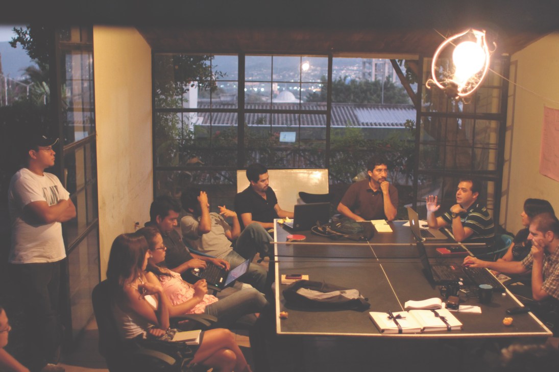 Carlos Dada leads a weekly editorial meeting with El Faro staff members seated around a table in an office with large windows overlooking San Salvador.