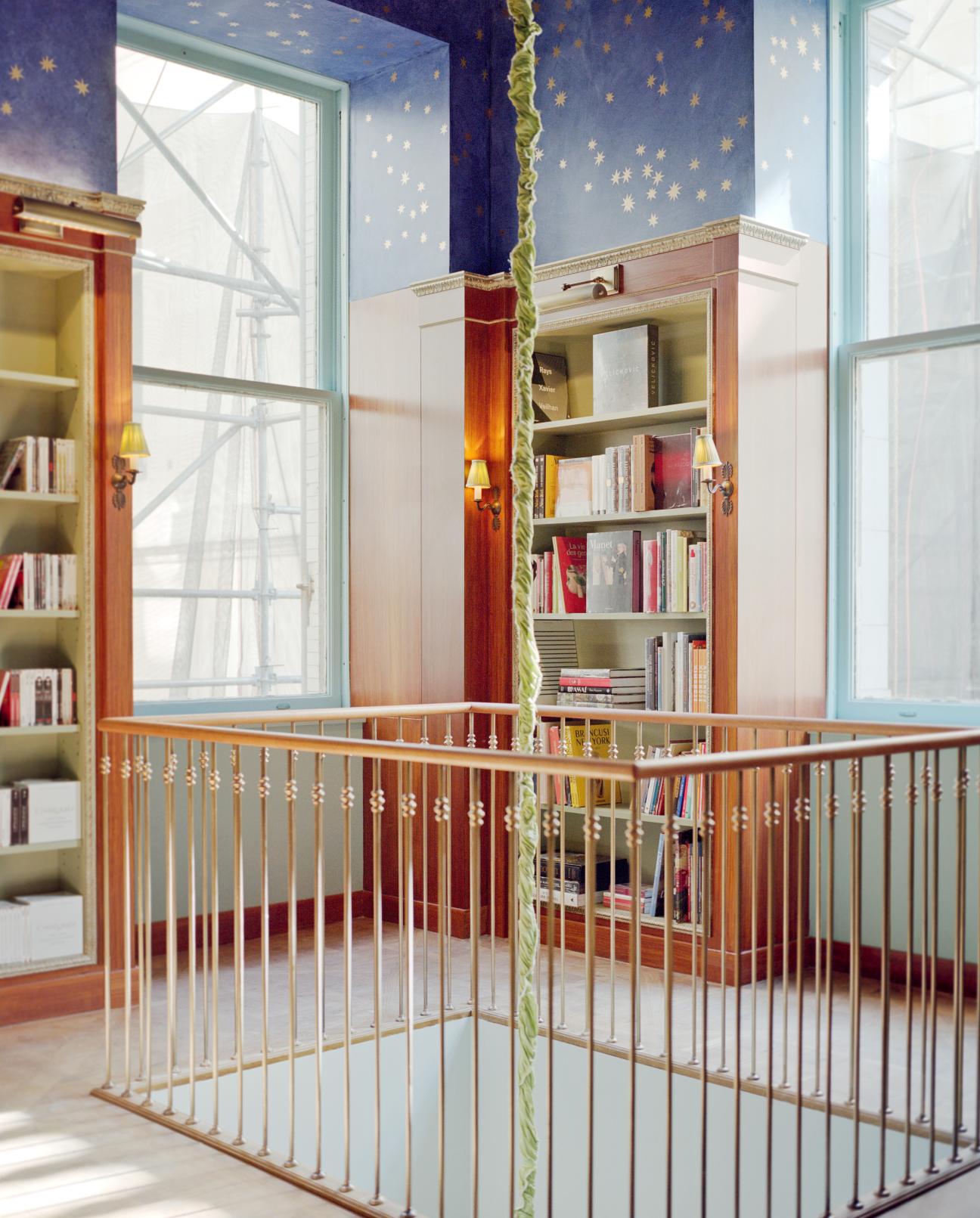 A multi-level bookshop interior with cream bookshelves, a starry blue ceiling fresco, and a brass railing dividing two floors.