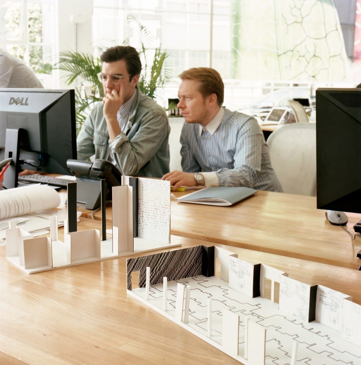 Two architects review designs at a desk with architectural models and computer monitors in a bright studio.
