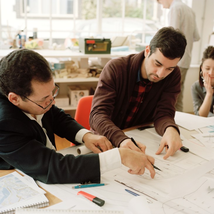 Architects reviewing design drawings at a desk in a bright studio workspace.