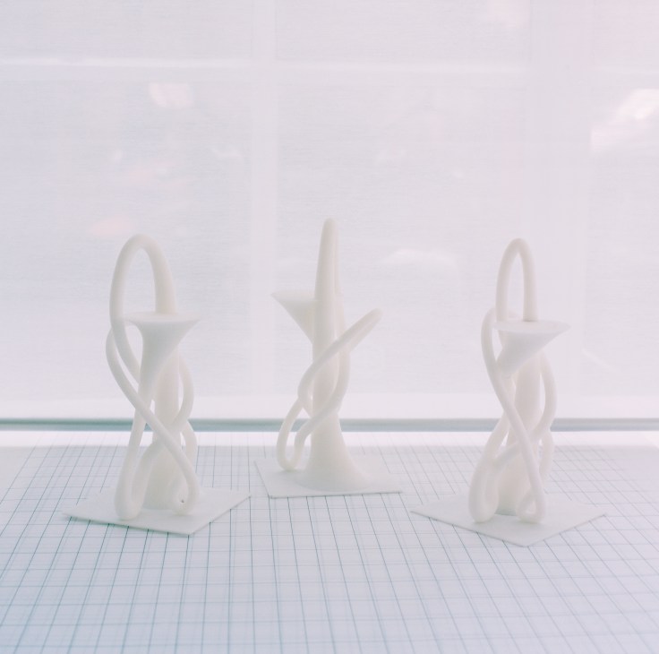Three white sculptural models with organic twisted forms displayed on a grid surface.