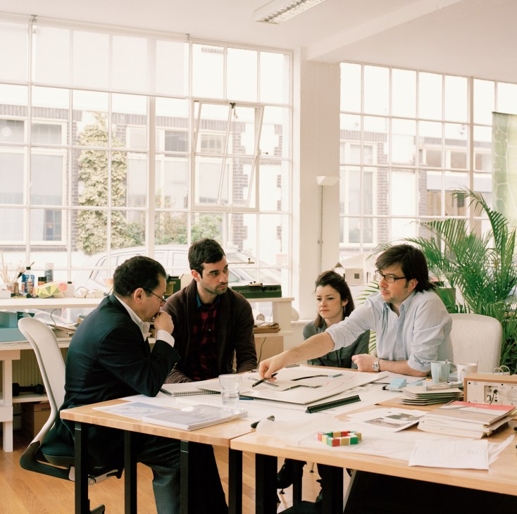 Four architects and designers in a bright studio meeting, reviewing designs and sketches at a wooden table.