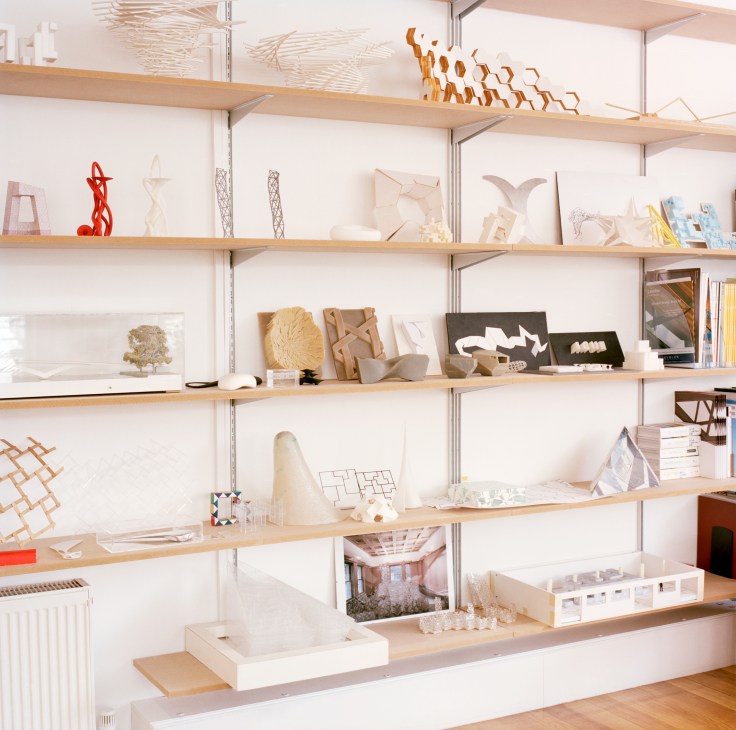 Shelving unit displaying architectural models, sketches, and design concepts in various materials and scales.