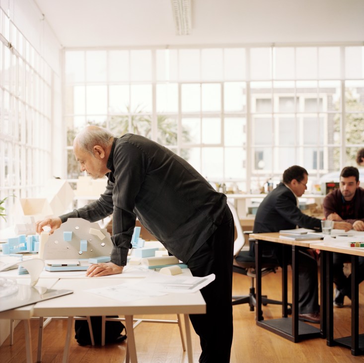 Cecil Balmond bends over a white table examining architectural models in a bright studio space.
