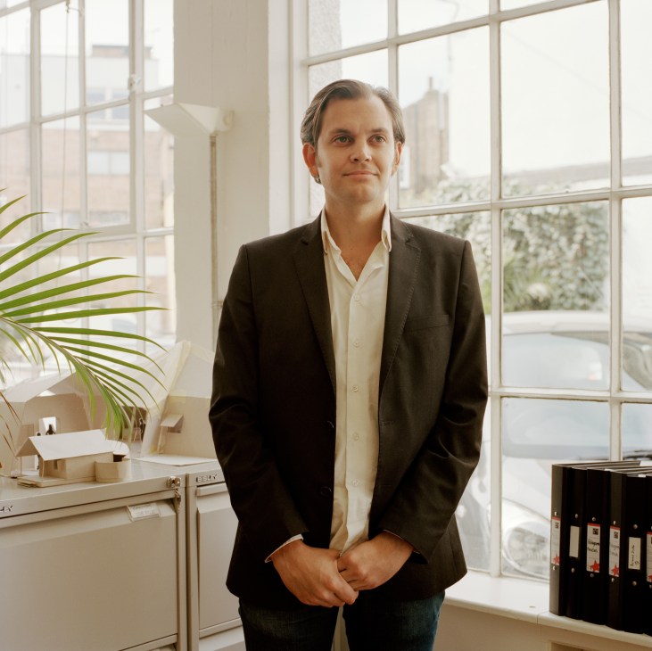 John Balmond stands in a bright office with large windows, wearing a dark blazer and white shirt.