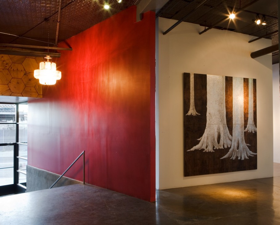 Modern art gallery interior with bright red wall and artwork featuring tree imagery on cream-colored wall.