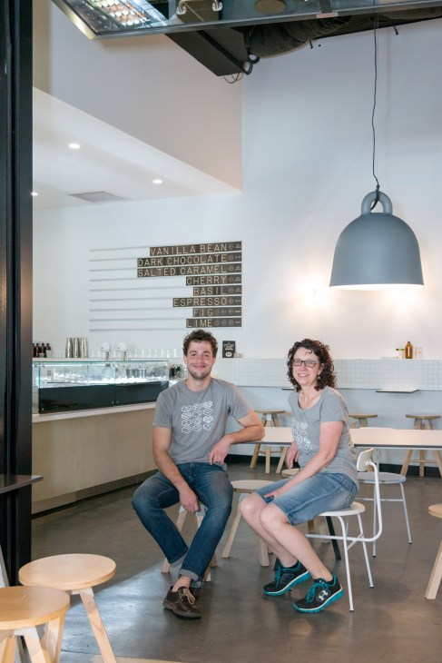 Two staff members sitting inside Frugii Ice Creamery with a menu board and pendant light visible.