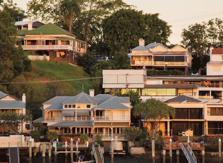 Waterfront houses with wooden decks and jetties along the Brisbane River at sunset.