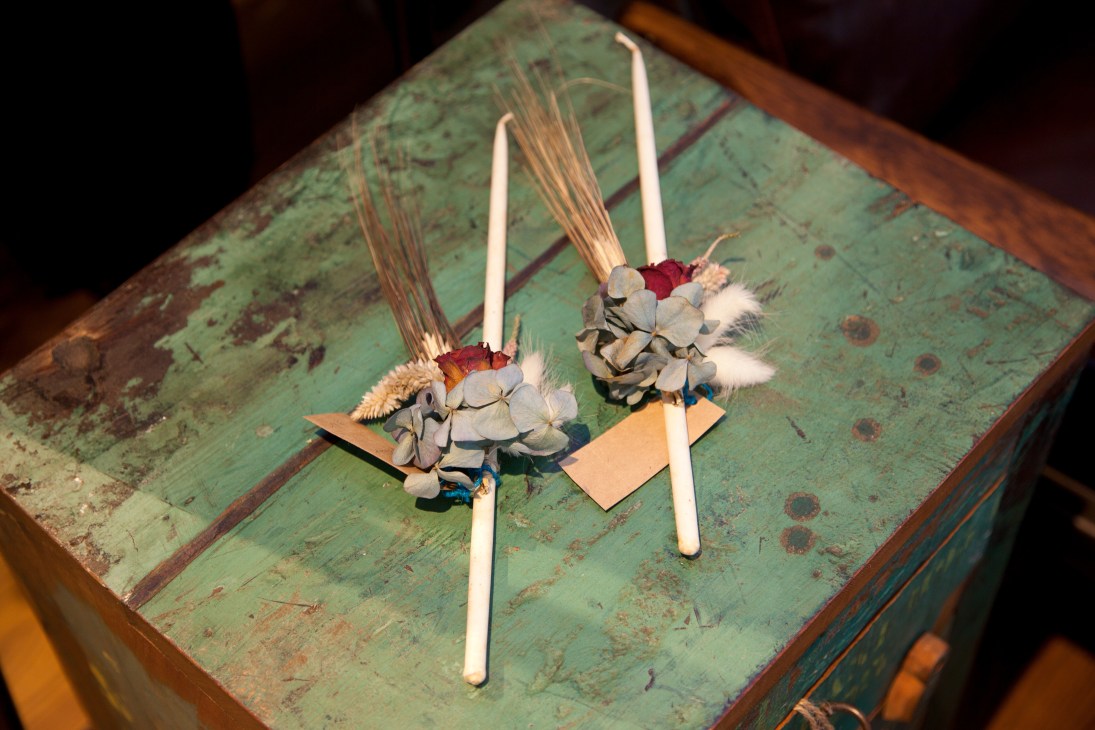 Two white candles with dried flower and grass arrangements on a weathered green wooden surface.