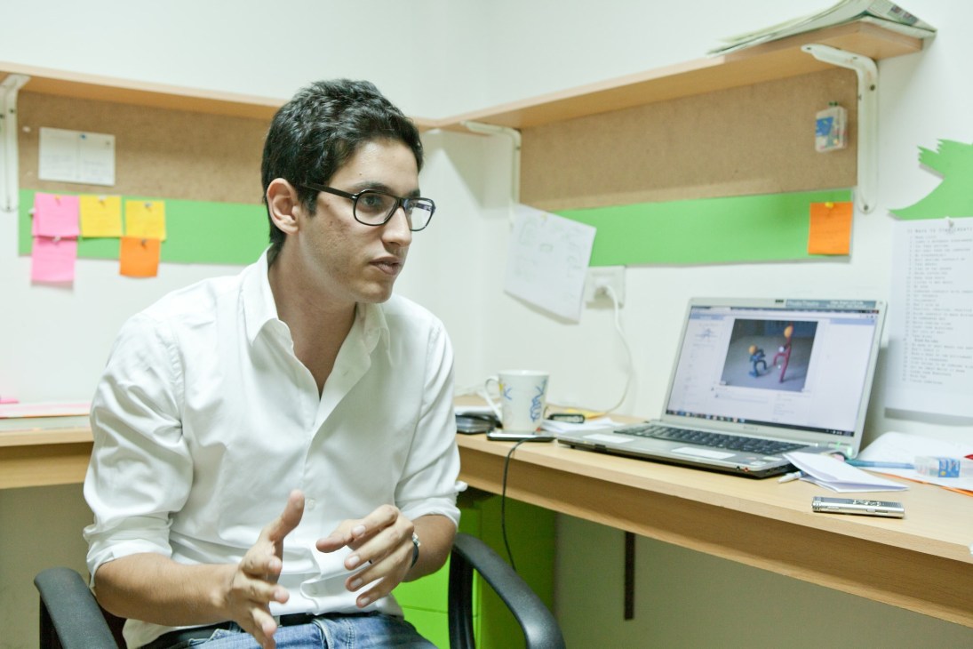 Bassem Elhady, a young man wearing glasses and a white shirt, sits at a desk with a laptop in his office, gesturing while speaking.