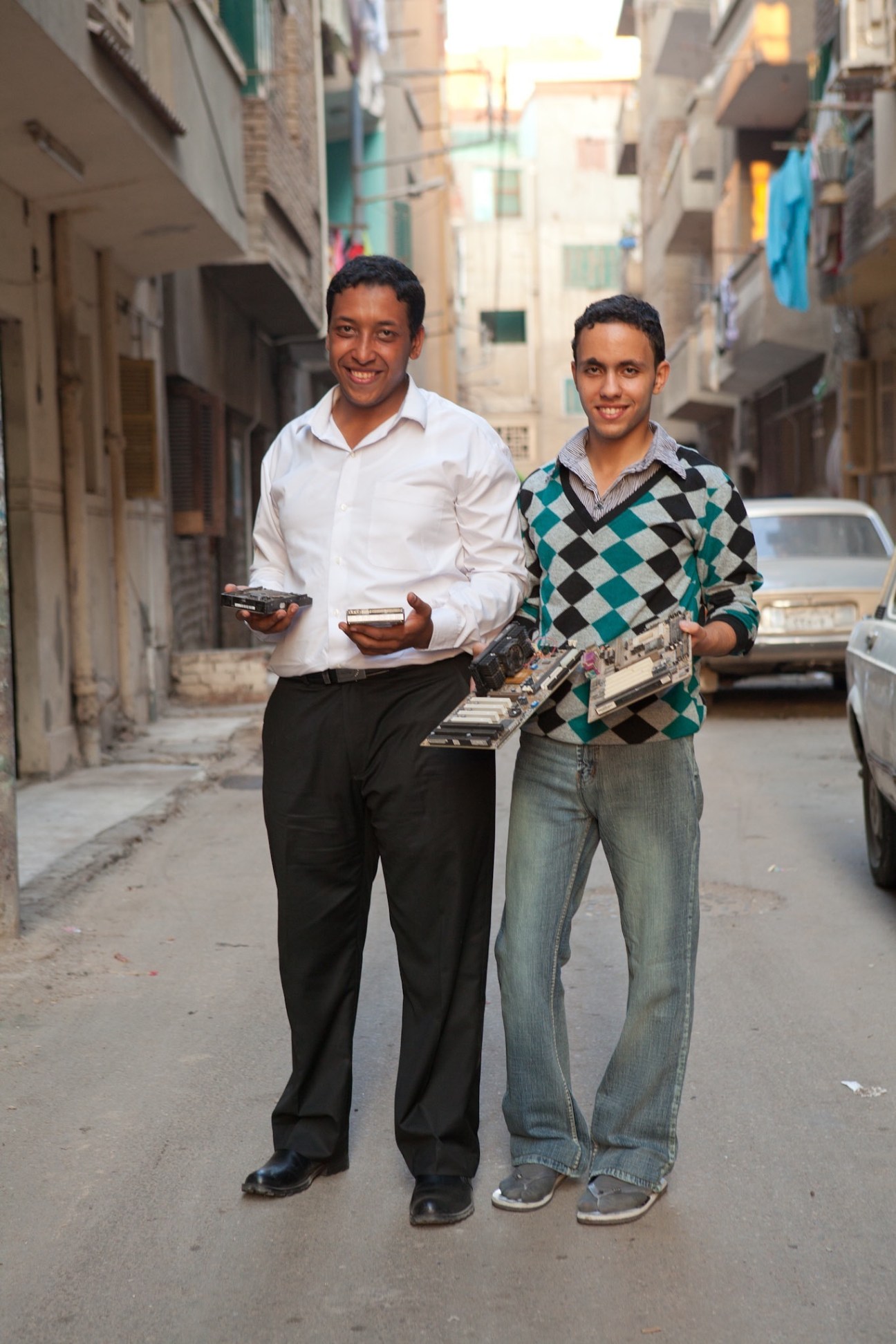 Two young men holding electronic components stand smiling in a narrow Cairo street lined with apartment buildings.