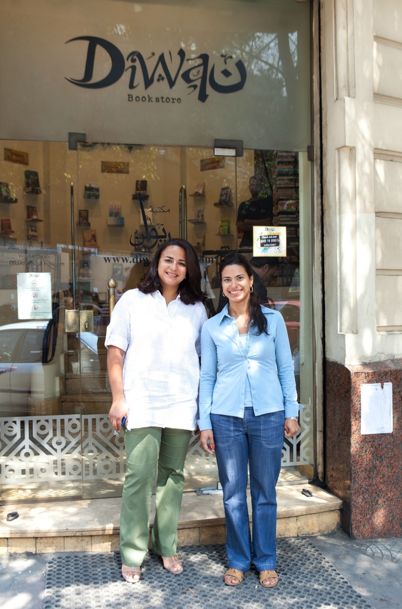Two women stand smiling outside Diwan Bookstore, with book displays visible through the window behind them.