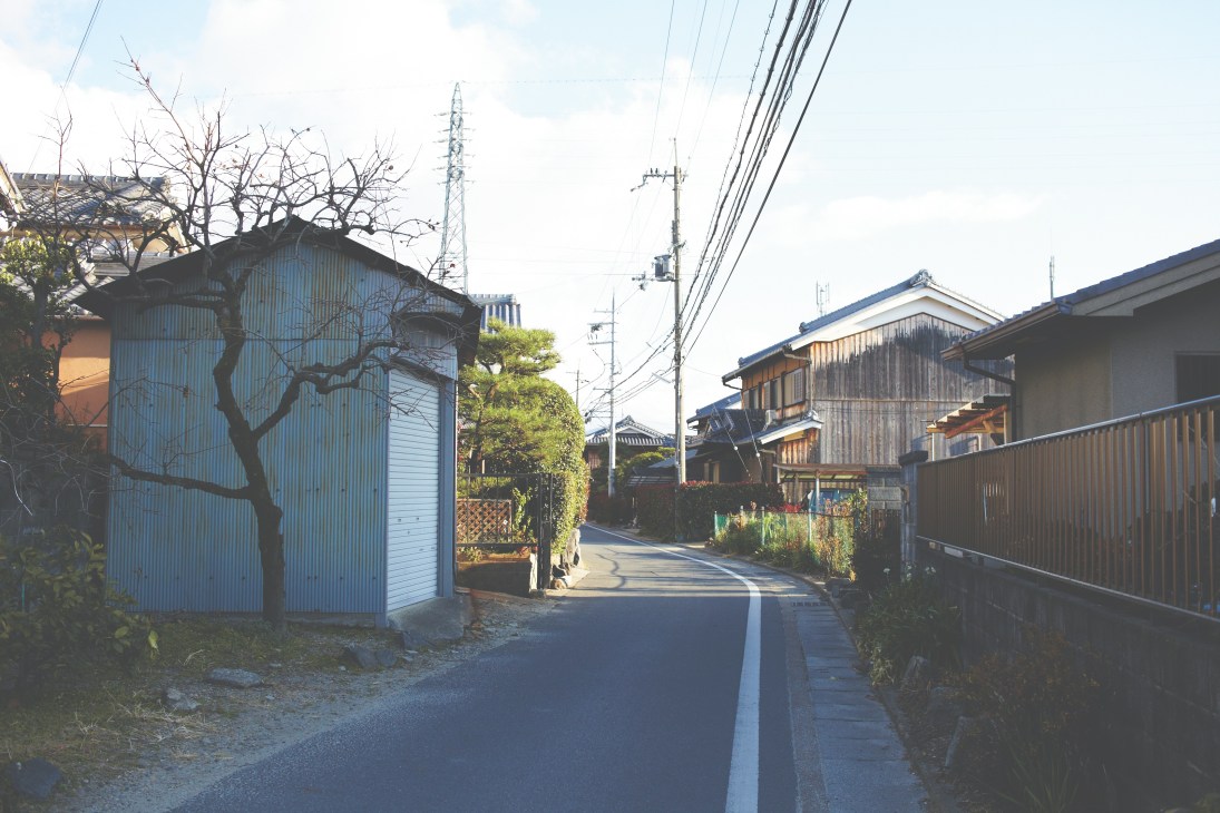 A quiet residential street in northern Kyoto lined with traditional wooden houses and corrugated metal structures, with power lines overhead.